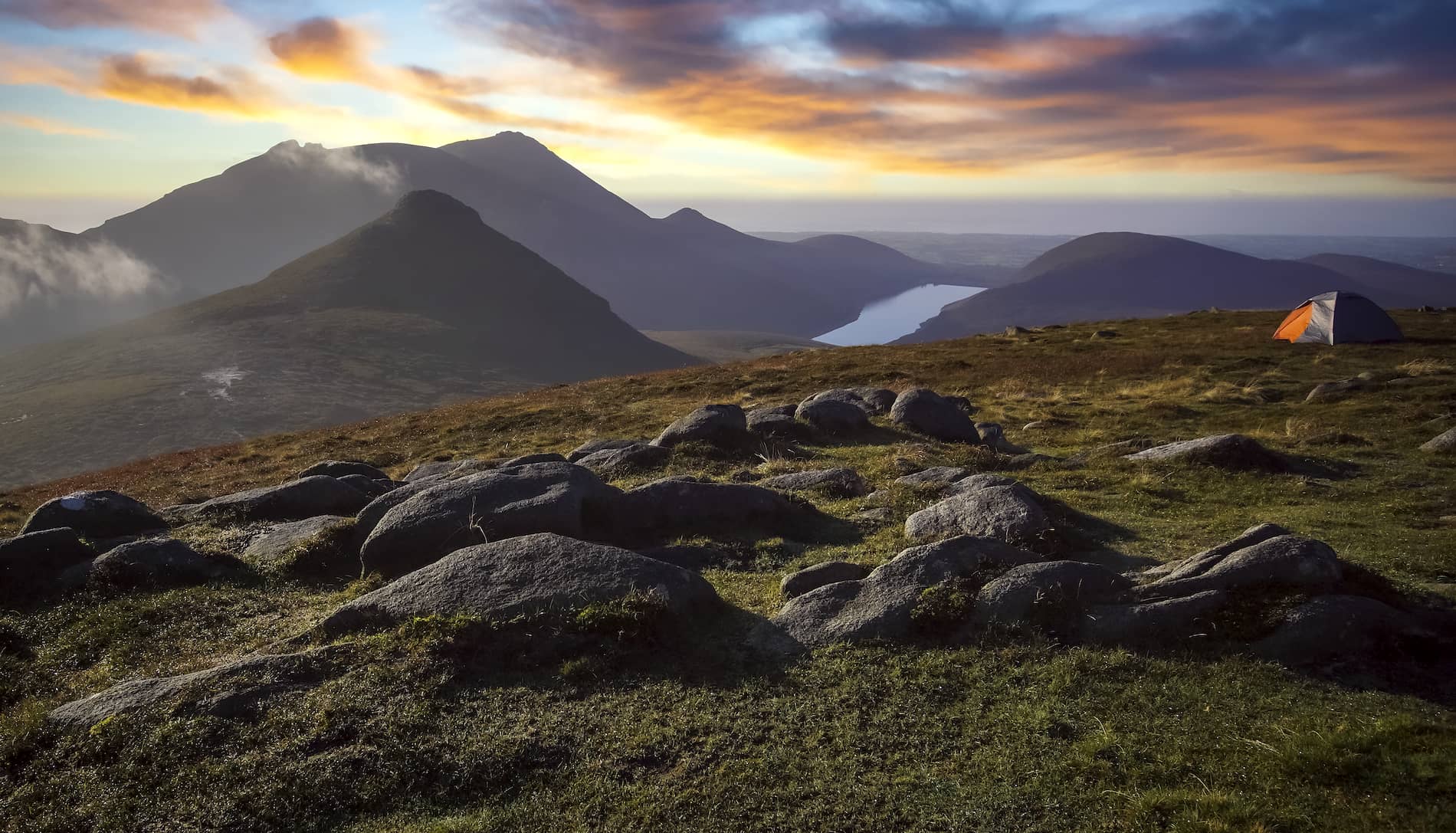 Sunrise over the Mourne Mountains in County Down with rocky peaks, a lakeside valley and a tent overlooking the landscape.