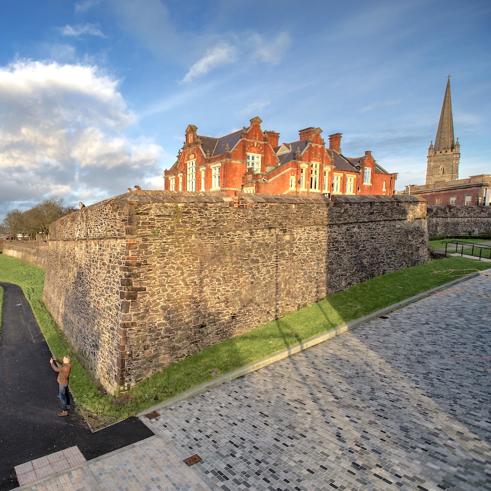 Die historischen Stadtmauern von Derry~Londonderry neben roten Backsteingebäuden und dem Kirchturm.