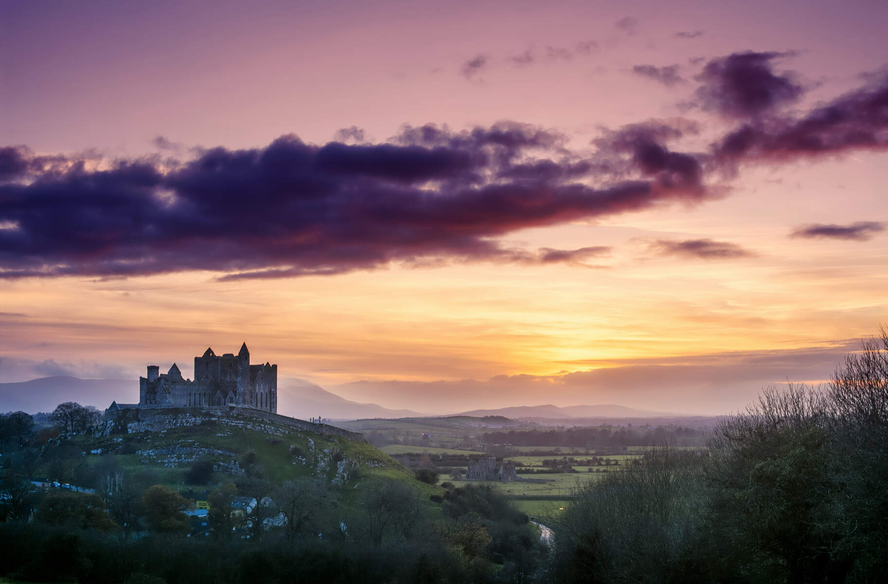 The Rock of Cashel silhouetted against a purple and orange sunset over County Tipperary.