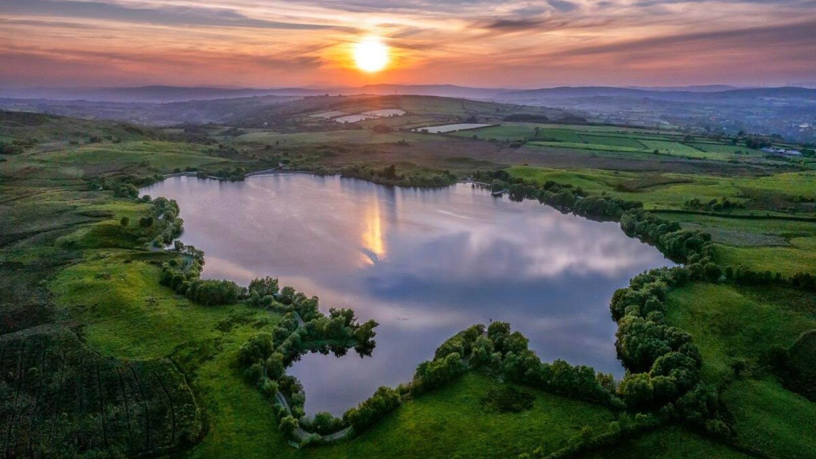 Sunset over Moorlough Lake, County Tyrone, reflected in calm water surrounded by green farmland.