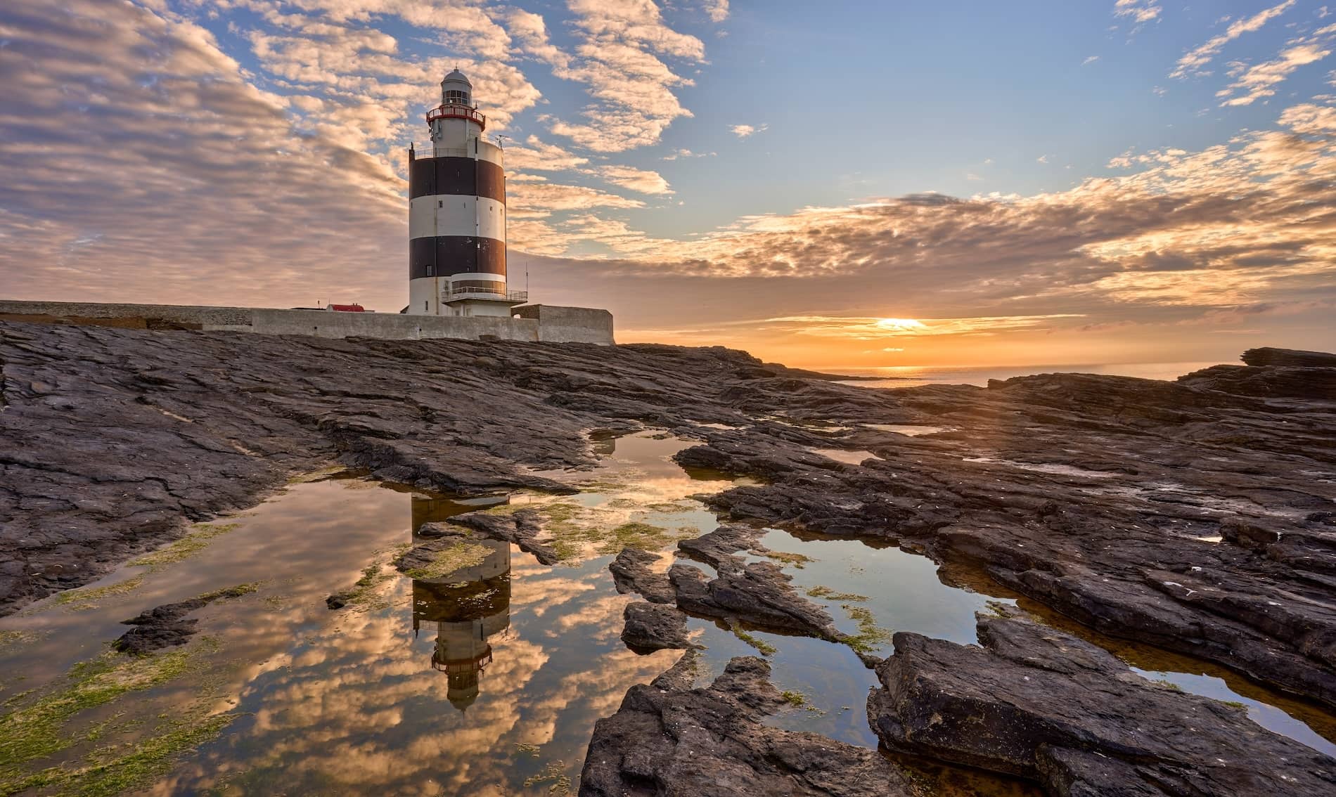 Hook Head Lighthouse in county Waterford weerspiegeld in rotsachtige branding bij zonsondergang onder een dramatische lucht.