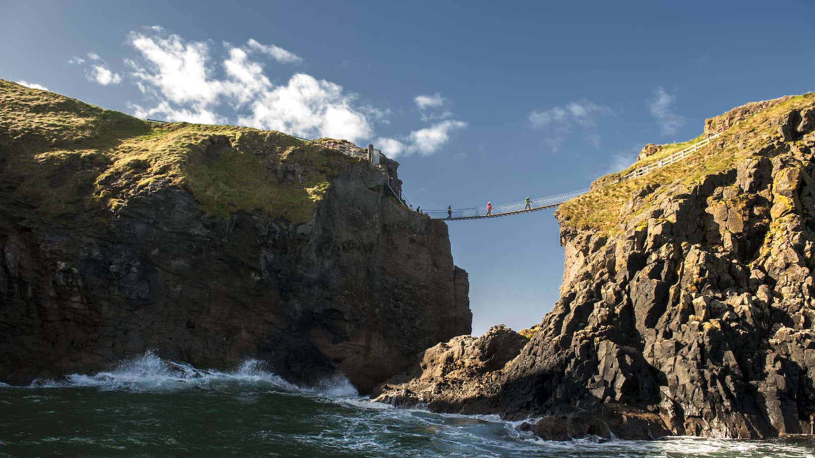 Visitors crossing the Carrick-a-Rede Rope Bridge between rugged sea cliffs on the Antrim Coast.