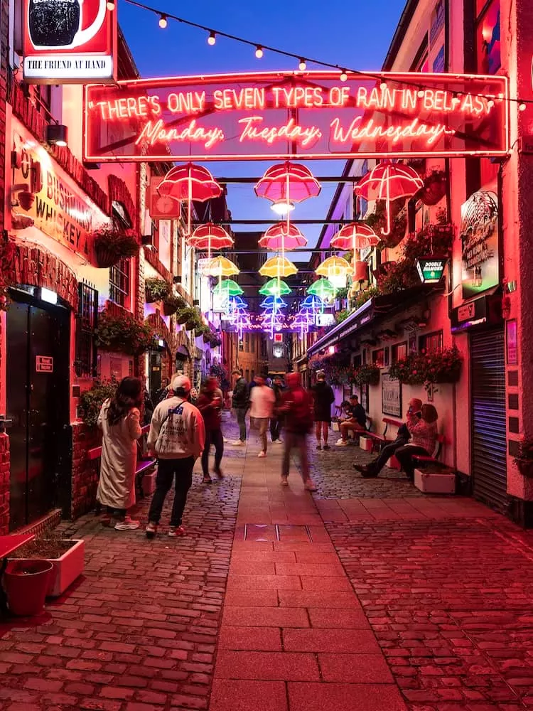 Handelsgericht in Belfast bei Nacht, leuchtend mit Neonschildern und farbenfrohen Schirmlichtern.