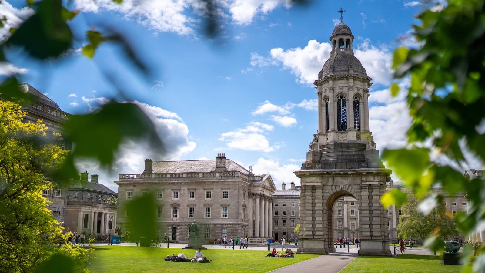 Students relaxing on the green at Trinity College Dublin beneath the Campanile on a sunny day.