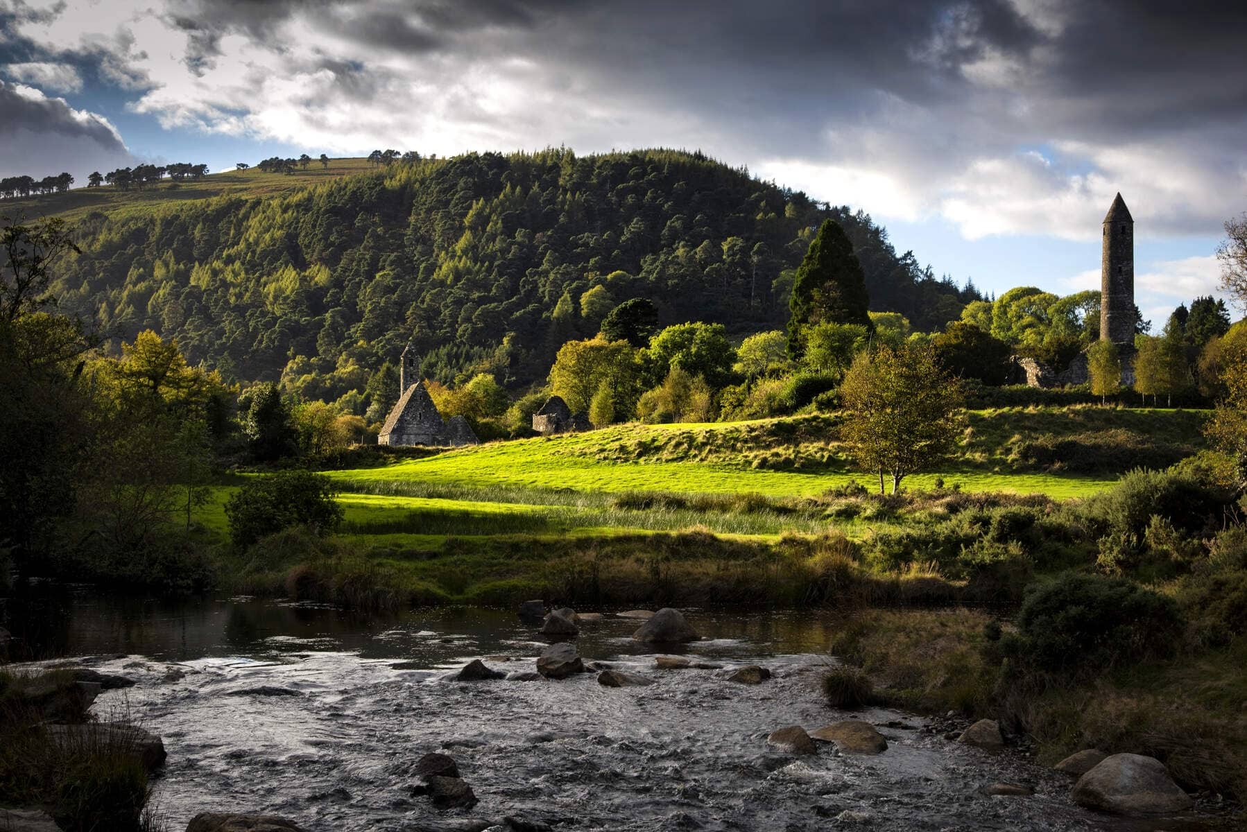Sunlight breaks through clouds over Glendalough's monastic ruins and round tower, nestled in a lush green valley by a flowing stream.