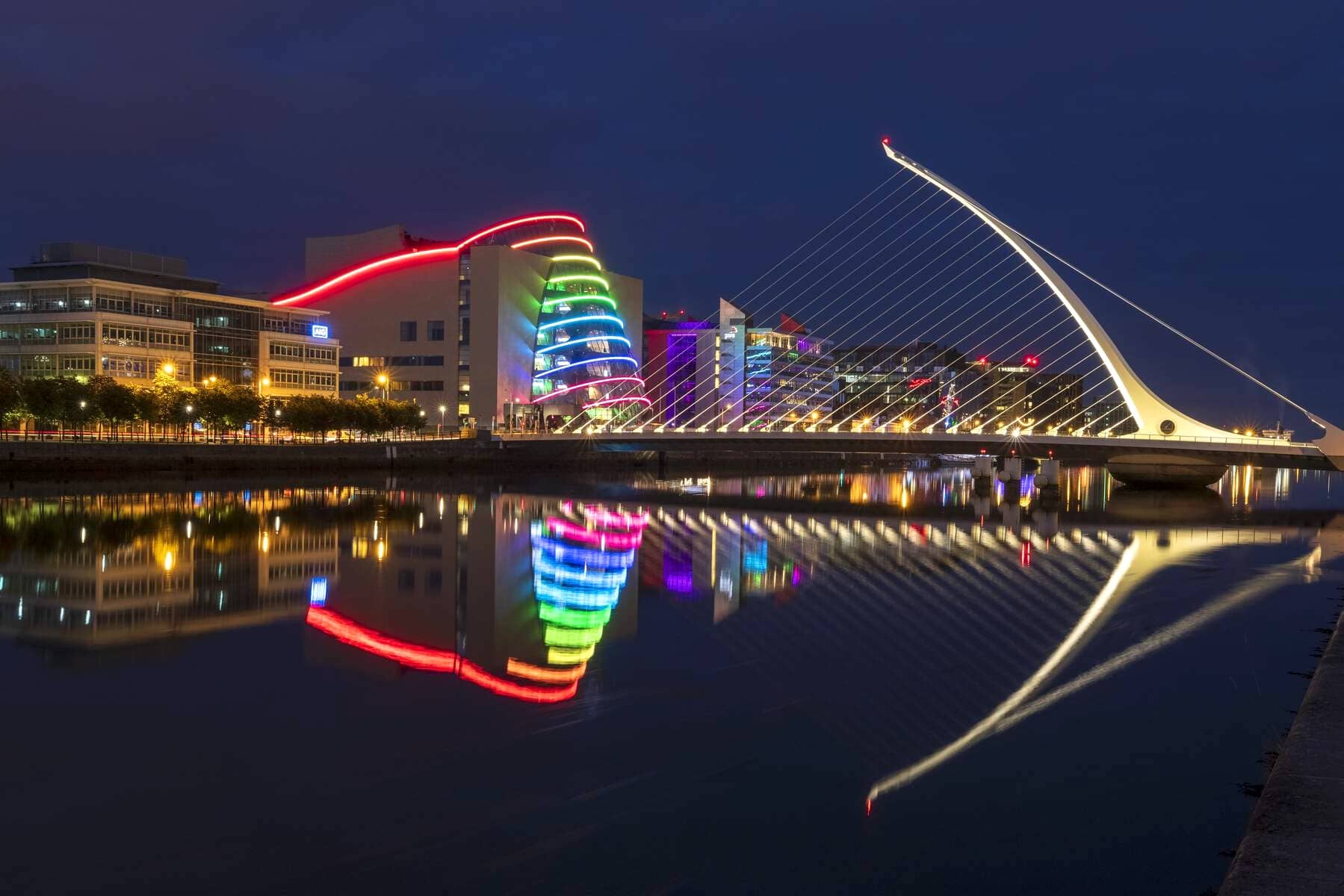 Colourfully lit Convention Centre and Samuel Beckett Bridge reflected in the River Liffey at night.