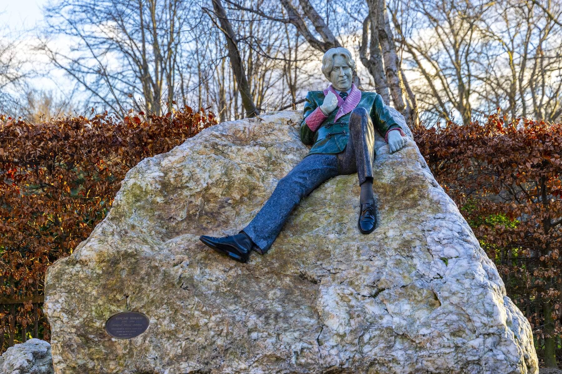 Statua colorata di Oscar Wilde sdraiato su una roccia, circondato da alberi spogli in inverno.