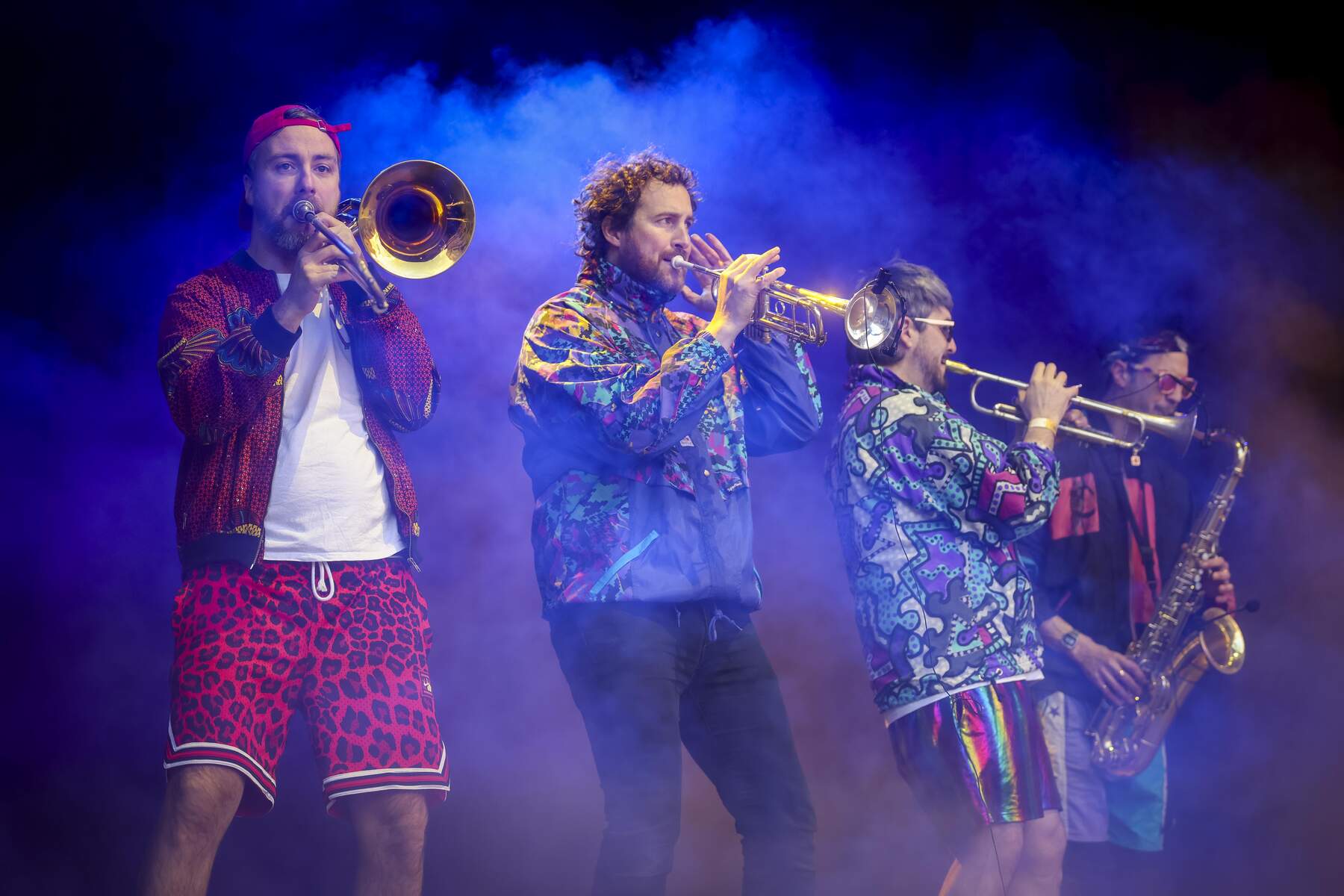 Musicians in vibrant outfits play brass instruments on stage, surrounded by dramatic purple and orange stage lighting.
