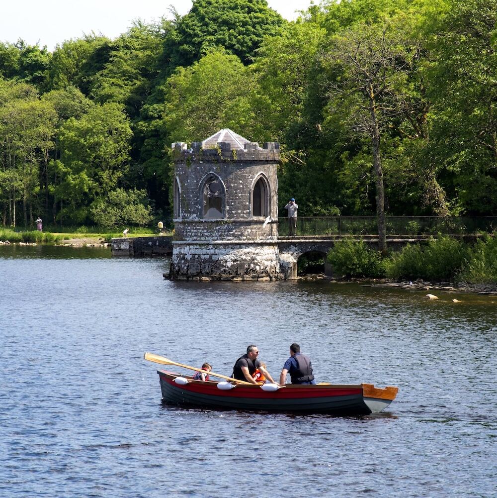 A family rows a boat past a stone folly tower on Lough Key, surrounded by lush green woodland in County Roscommon.