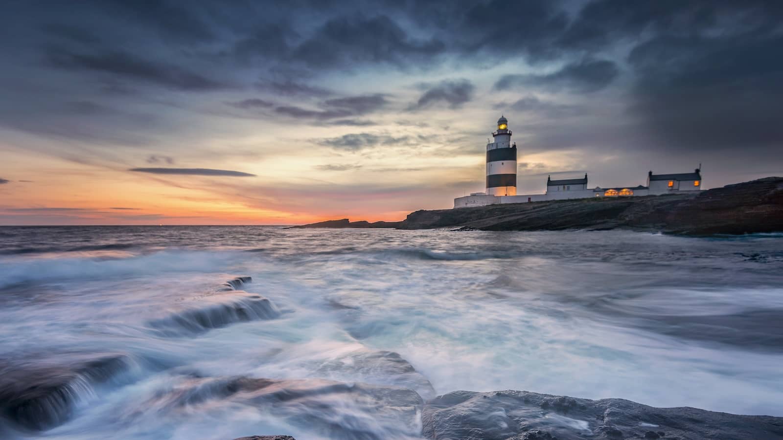 Hook Head Lighthouse standing on a rocky coastline of County Wexford as waves crash against the shore.