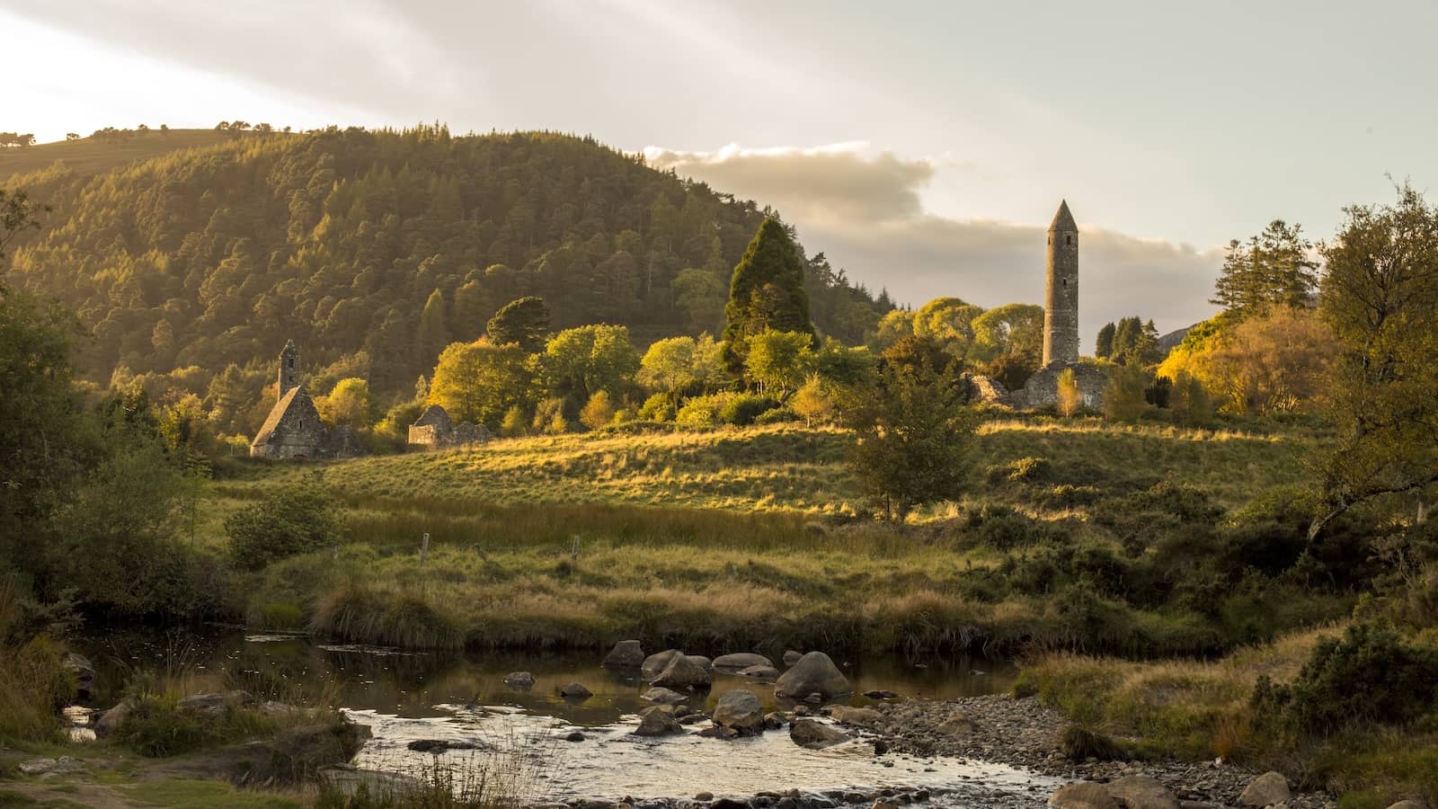 Round tower and monastic ruins at Glendalough, County Wicklow, set among wooded hills and a flowing stream.