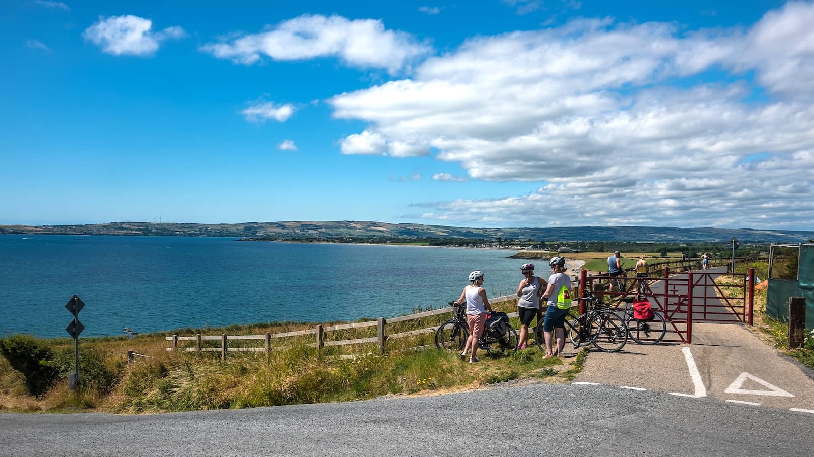 Cyclists on the Waterford Greenway stopping to admire coastal views across a calm bay under a bright blue sky.