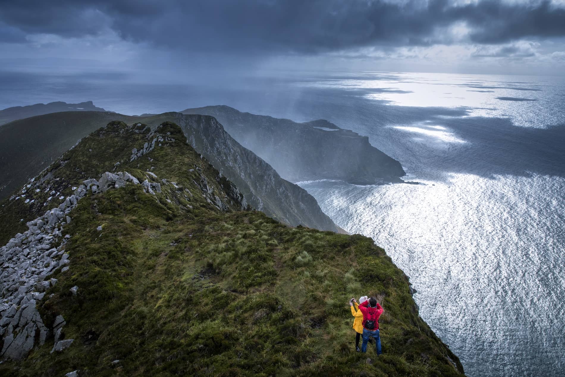 Two hikers at the edge of the towering Slieve League Cliffs in County Donegal with dramatic coastal views and moody clouds overhead.