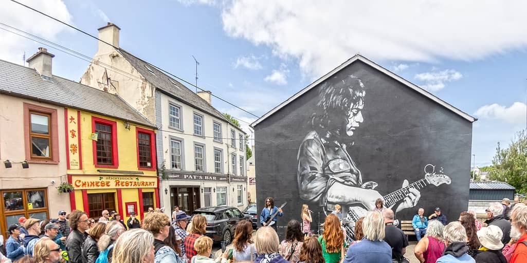 Crowd gathered in front of a large monochrome mural of Rory Gallagher playing guitar during his namesake festival in Ballyshannon, Donegal.