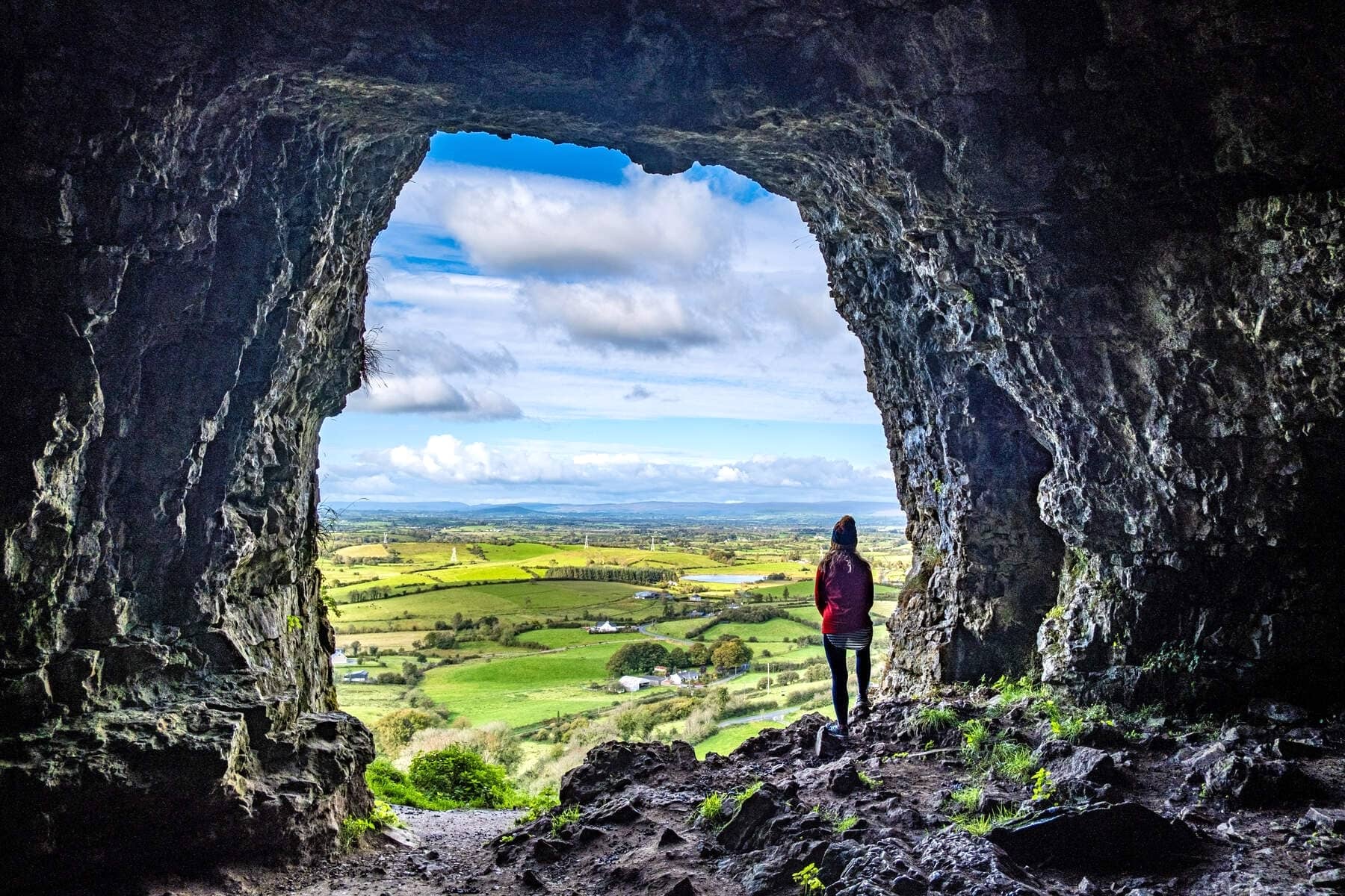 Personne debout à l'entrée des grottes rocheuses de Keash, surplombant une vallée verdoyante et des terres agricoles en mosaïque dans le comté de Sligo.