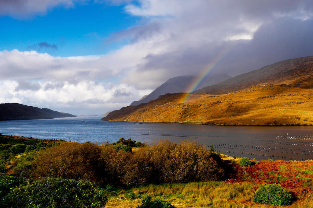 Rainbow over Killary Harbour in Connemara with autumn-coloured hills and calm blue water under a bright sky.