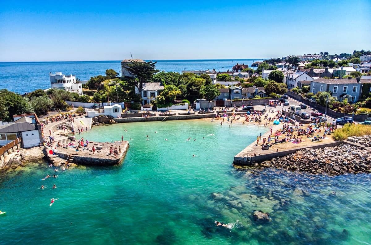 Forty Foot bathing area in Sandycove, County Dublin, with swimmers in turquoise sea beside seaside homes.