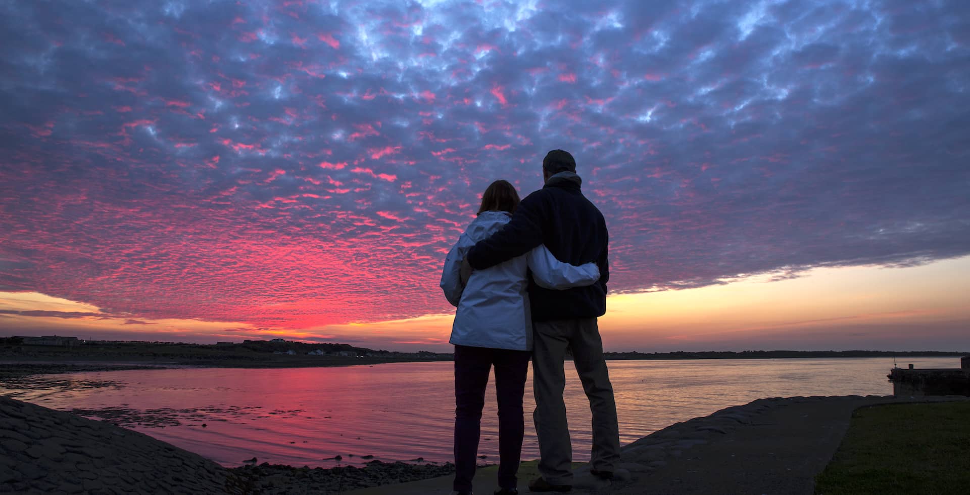 Couple debout, bras dessus bras dessous, admirant un coucher de soleil rose et violet éclatant sur les eaux calmes au large de Burr Point, dans le comté de Down.