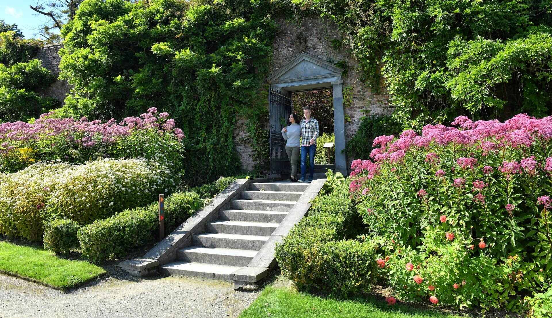 Pareja caminando bajo un arco de piedra rodeado de vibrantes flores de verano en los jardines Mount Congreve, condado de Waterford.