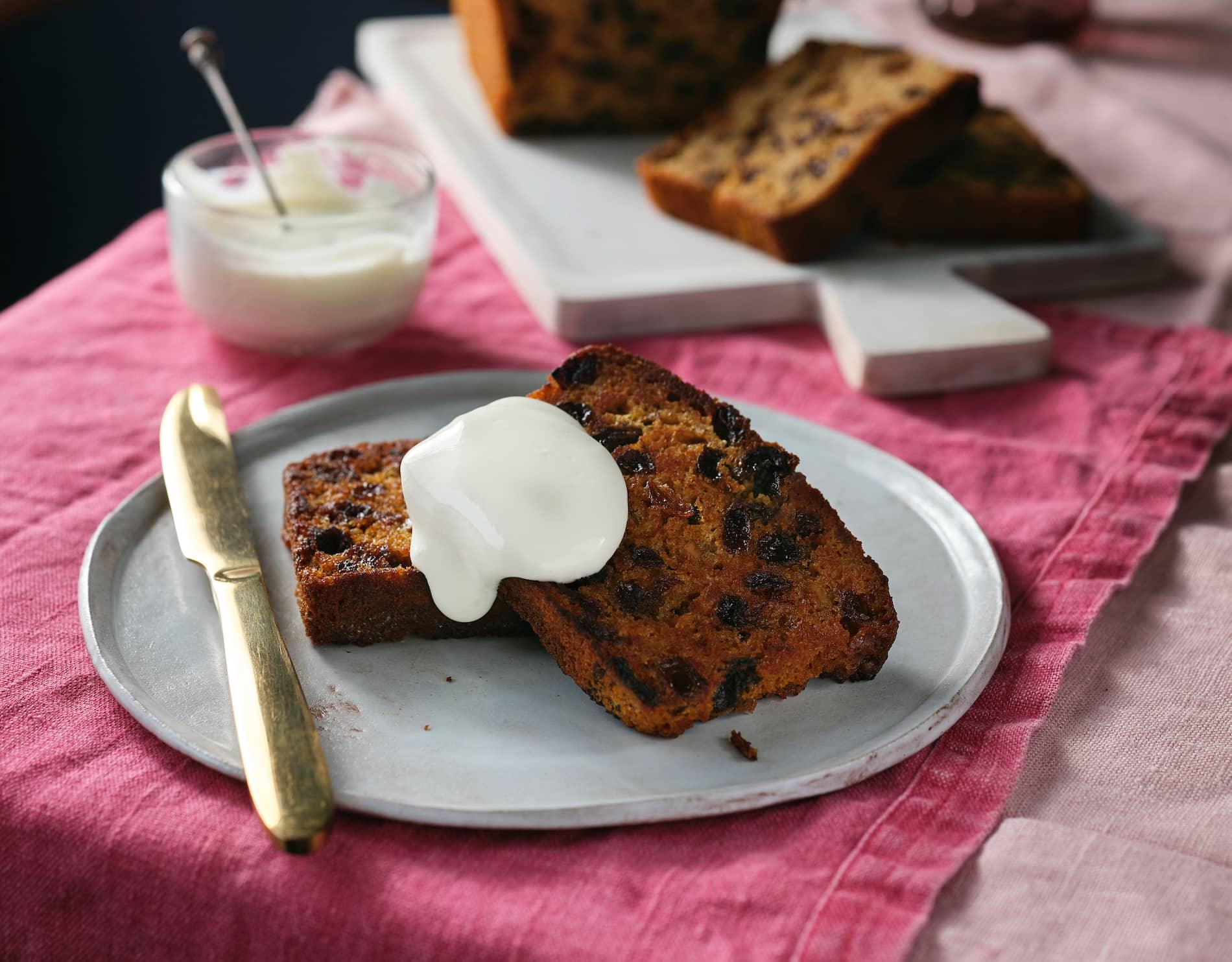 Slices of traditional Irish barmbrack topped with cream on a ceramic plate, with a loaf and more slices in the background.