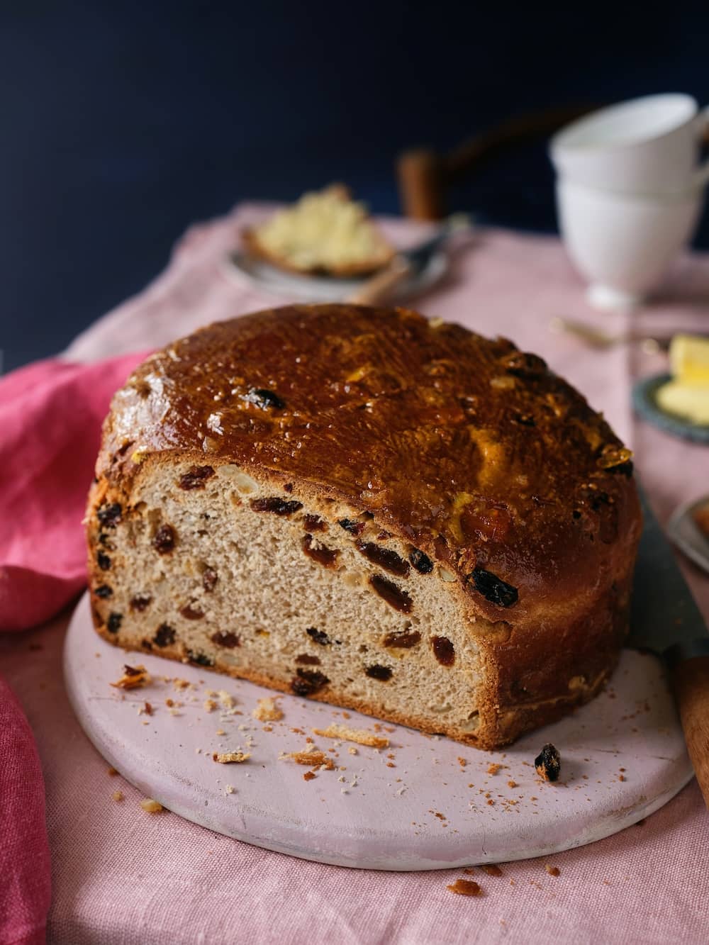 A sliced loaf of traditional Irish barmbrack filled with dried fruit, served on a pink board with butter and tea.