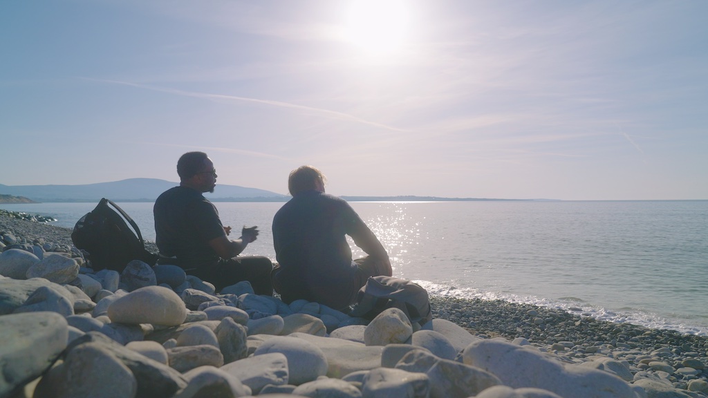 Babatunde Aléshé and Finlay Christie sit on a rocky shore at Strandhill Beach, looking out at the calm sea under bright afternoon sun.