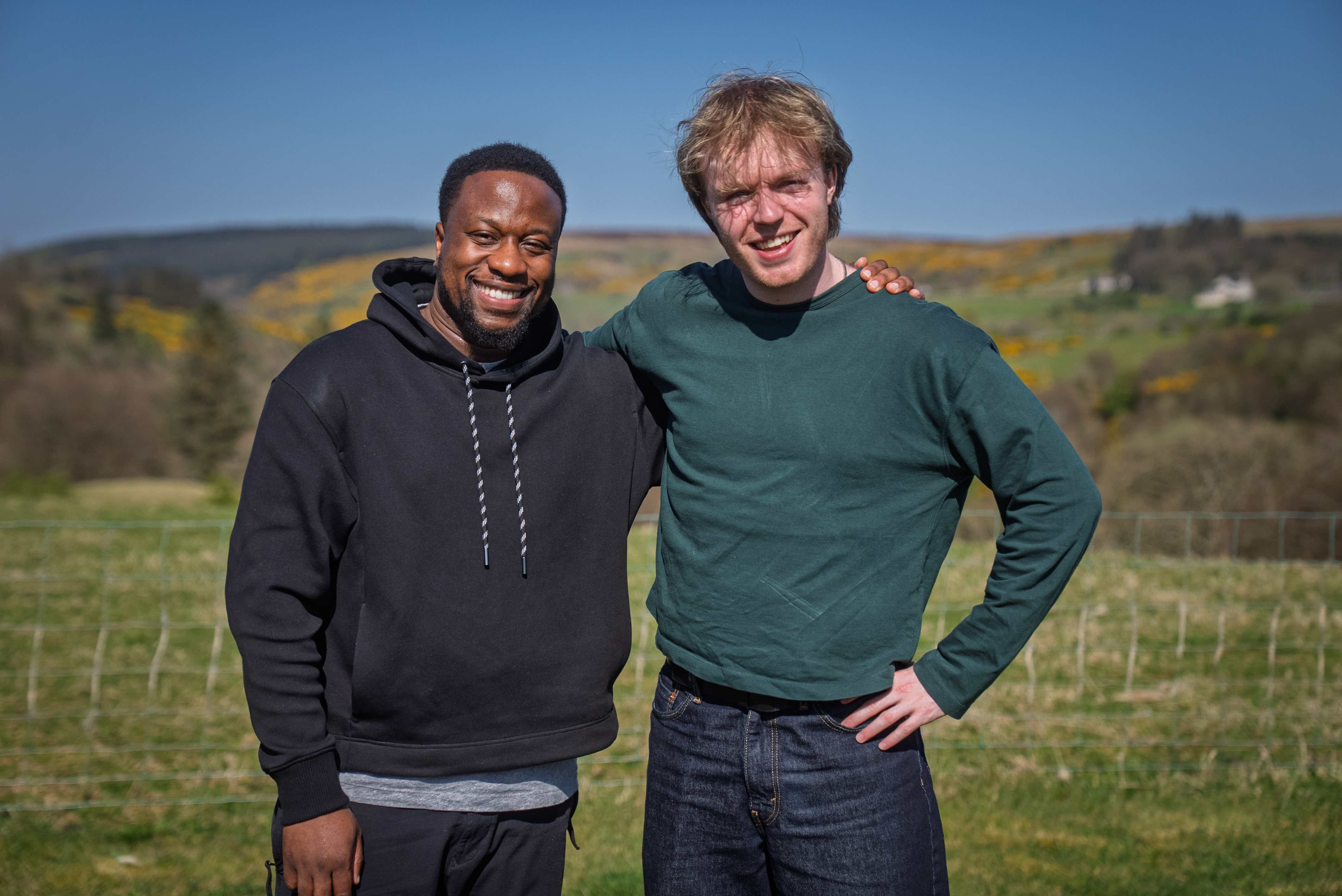 Babatunde Aléshé and Finlay Christie stand arm-in-arm outdoors, smiling warmly with green hills and gorse of Ireland's north coast in the background.