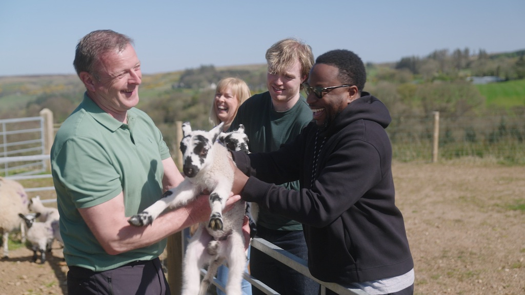 Babatunde Aléshé and Finlay Christie laugh as they help hold a young lamb at Dunfin Farm, joined by two smiling farmers on a sunny day.