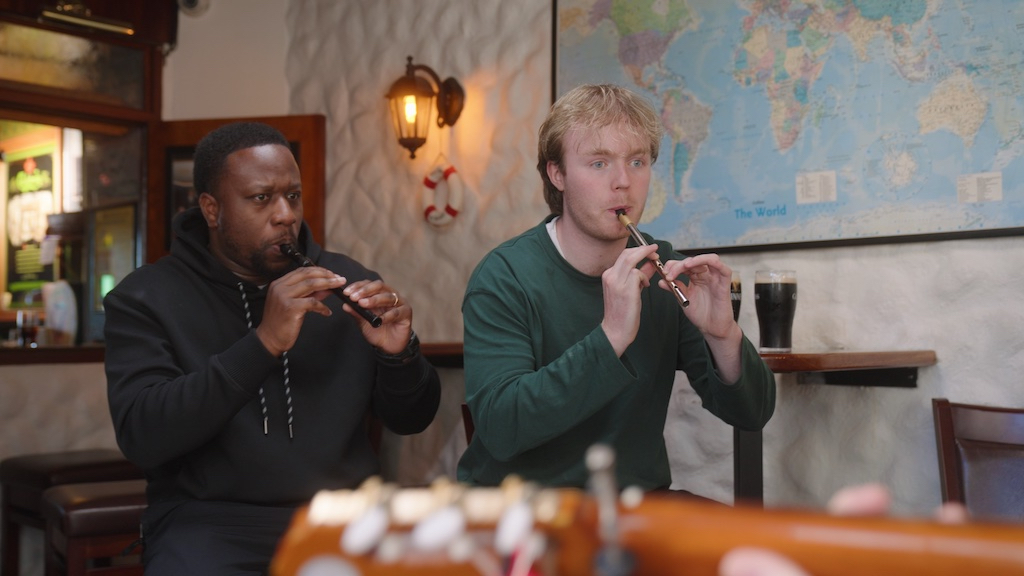 Babatunde Aléshé and Finlay Christie play tin whistles inside a cosy pub, surrounded by musical instruments and pints.