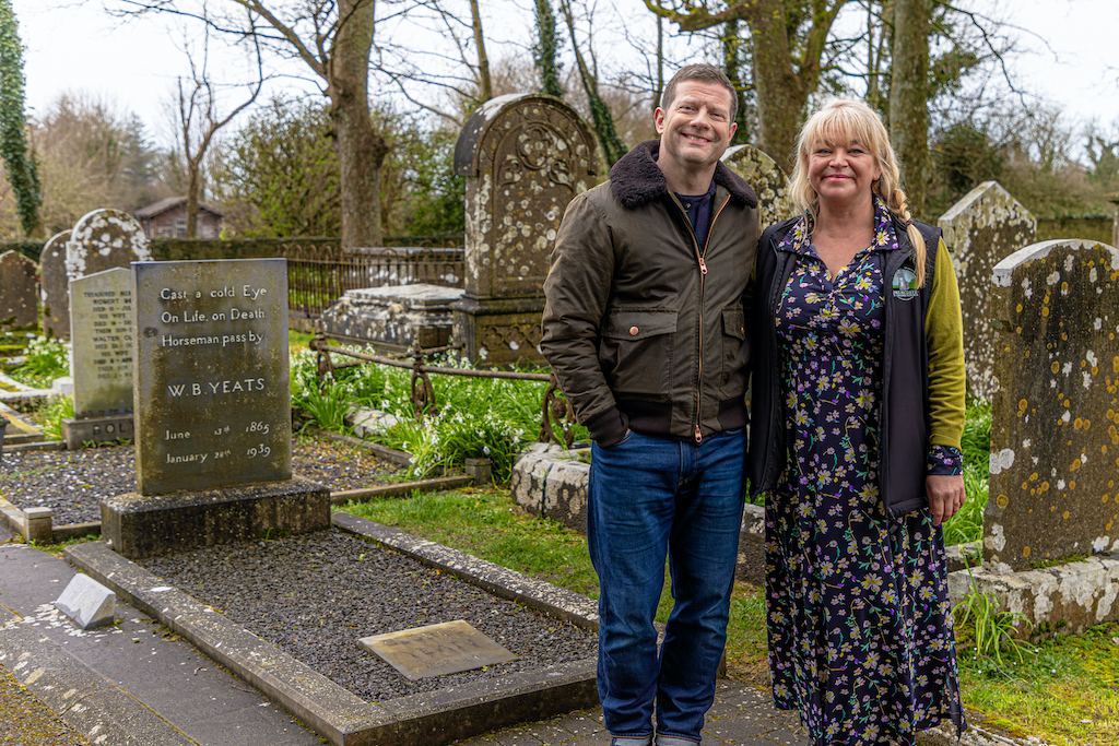 Dermot O'Leary and woman standing beside WB Yeats' grave in Sligo, surrounded by historic headstones in a quiet graveyard.