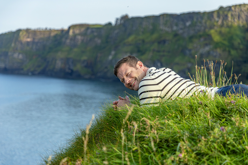 Dermot O'Leary lying on grass near Kinbane Castle cliffs, overlooking the sea and dramatic coastal scenery in Northern Ireland.