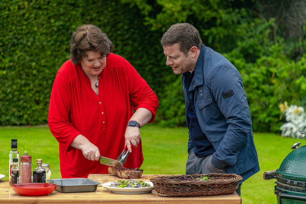 Chef Paula McIntyre slicing roast beef outdoors at Glenarm Castle, with Dermot O'Leary watching beside a barbecue and garden setting.