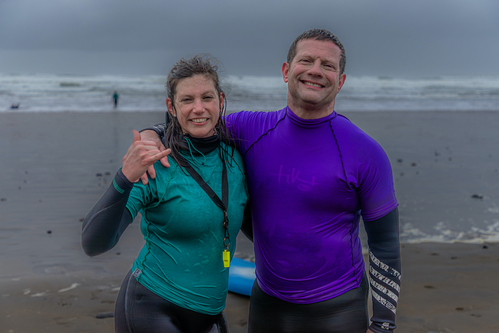 Dermot O'Leary and surfer in wetsuits smiling on Strandhill Beach after surfing, with waves and misty shoreline behind.