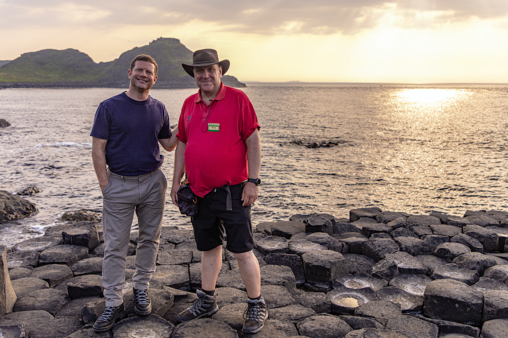 Dermot O'Leary and a storyteller standing on basalt columns at the Giant’s Causeway at sunset, with sea views and rugged coastline behind.