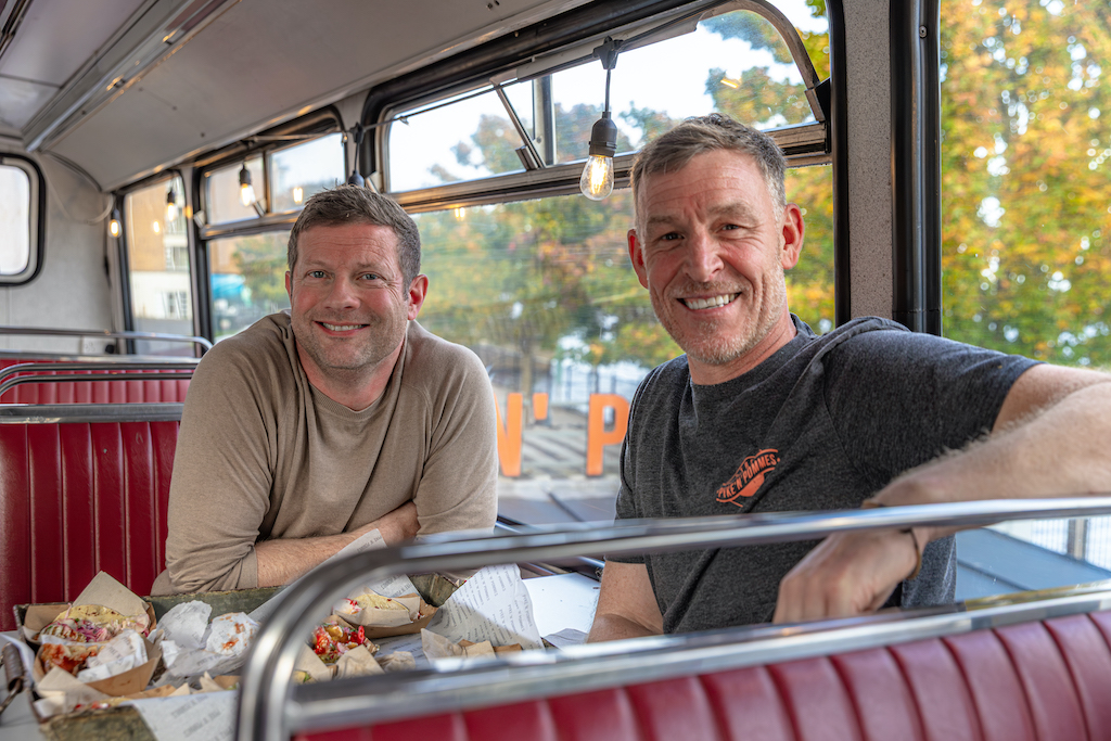 Dermot O'Leary and owner seated inside Pyke ’n’ Pommes in Derry~Londonderry, enjoying loaded chips at a retro bus-style dining booth.