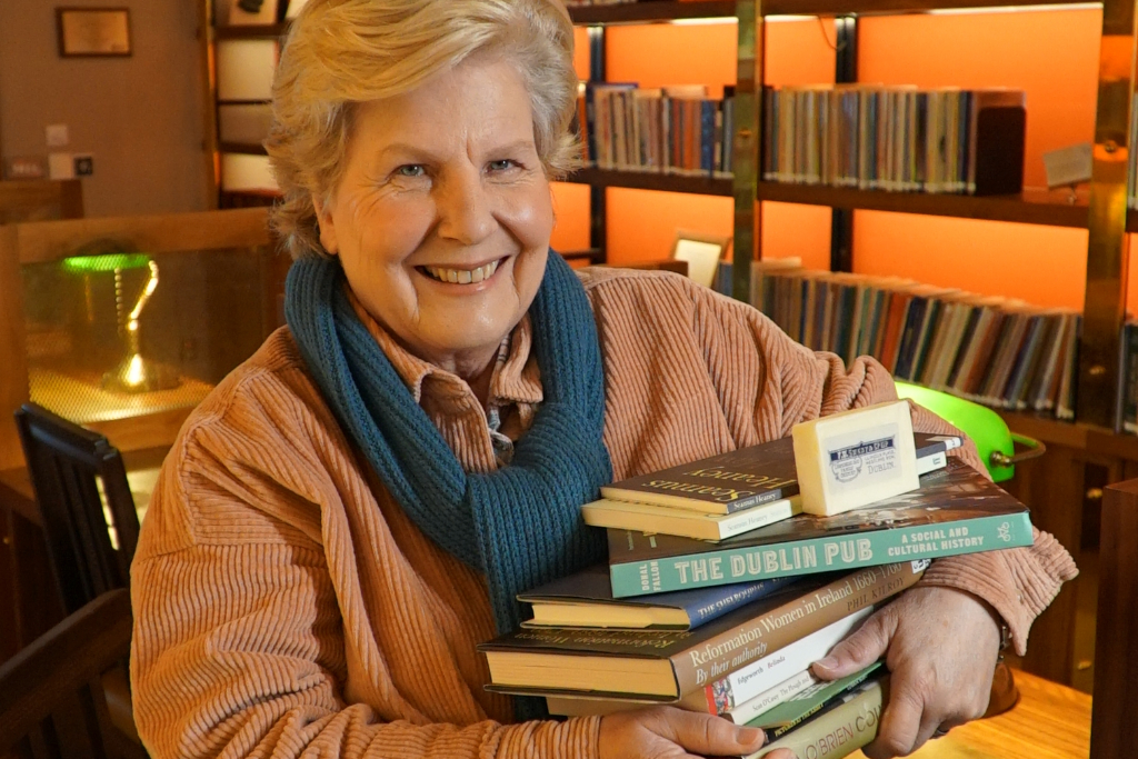 Sandi Toksvig smiles while holding a stack of books about Dublin and Irish history in a warmly lit, book-lined room.