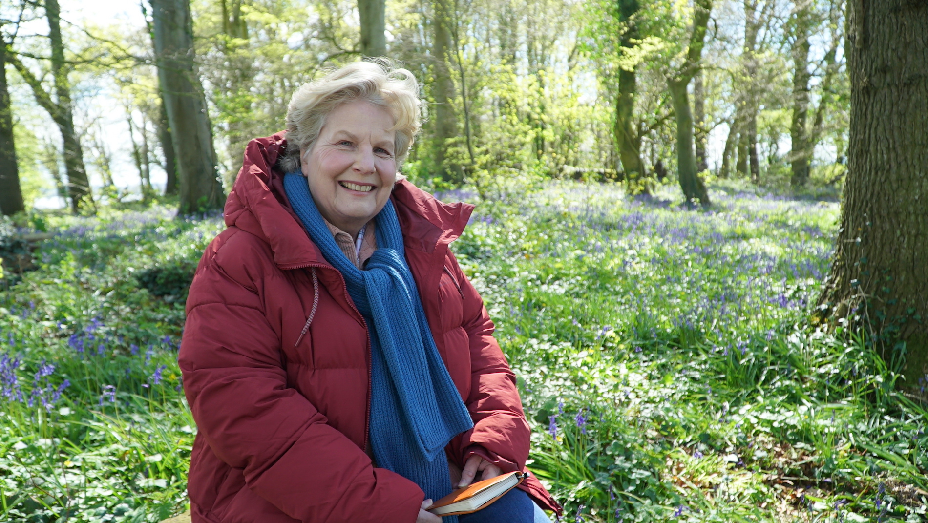 Sandi Toksvig smiles among bluebells in a sunlit woodland, wearing a red coat and blue scarf and holding a notebook.