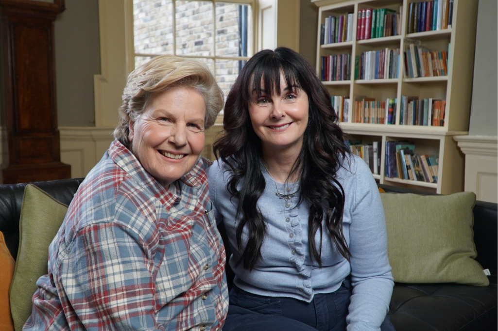 Sandi Toksvig and Marian Keyes smile together on a sofa, surrounded by shelves filled with books in a cosy room.