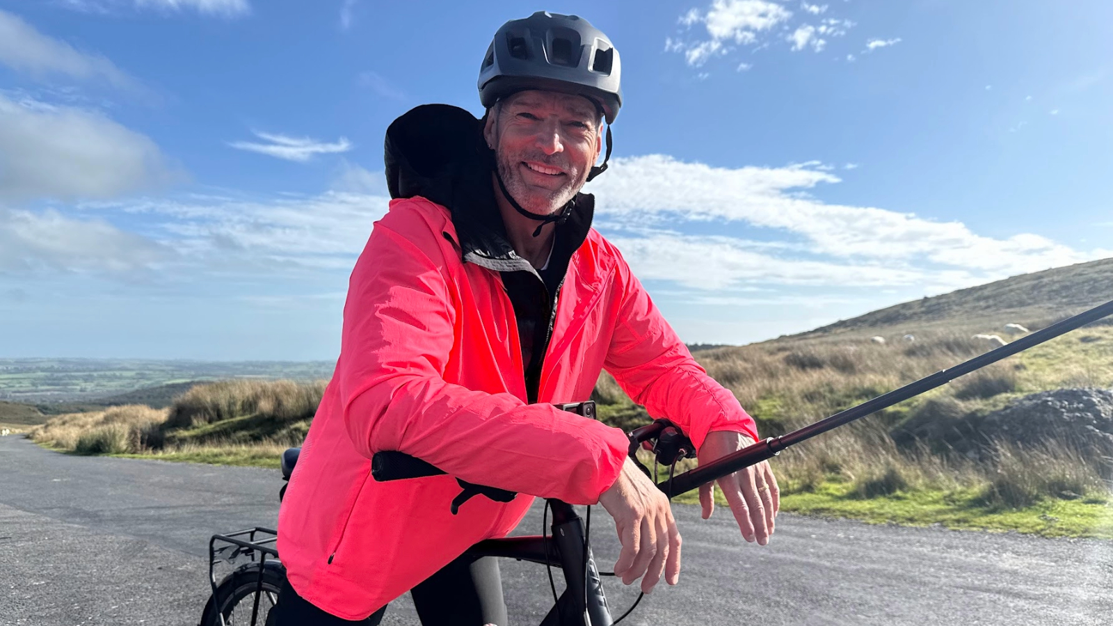 Fred Siriex resting on a bike on a rural road in the Comeragh Mountains, County Waterford, with rolling hills and blue sky behind.