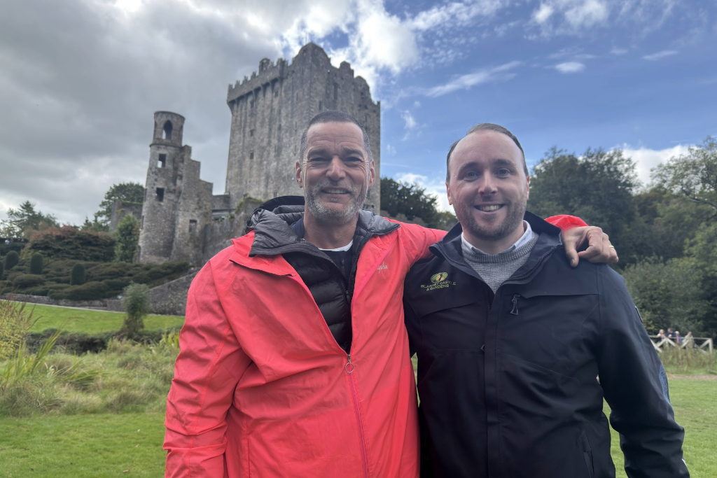 Fred and a tour guide smiling in front of Blarney Castle, with the medieval stone tower rising above landscaped gardens in County Cork.