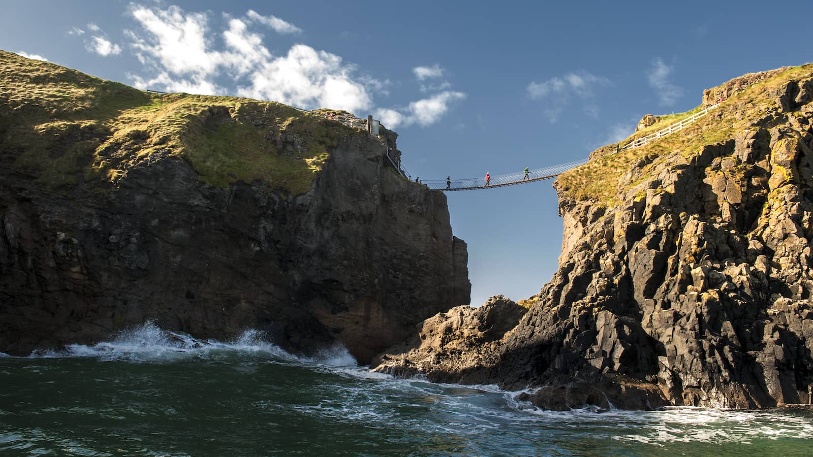 Visitors crossing the Carrick-a-Rede-rope bridge in County Antrim between rugged sea cliffs above crashing Atlantic waves.
