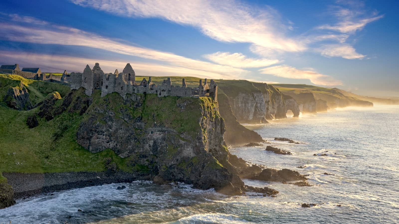 Ruins of Dunluce Castle, County Antrim, on a steep cliff above the Atlantic, with rugged coastline, sea arches and soft golden light at sunset.