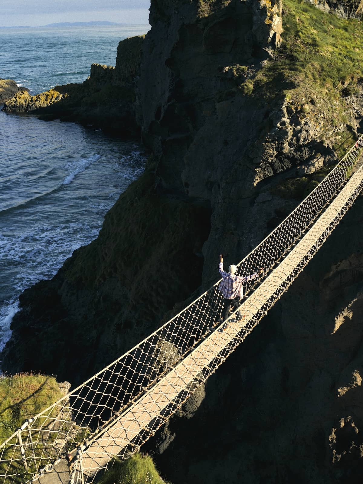 Person crossing the Carrick-a-Rede rope bridge above a rocky coastline with waves breaking below in County Antrim.