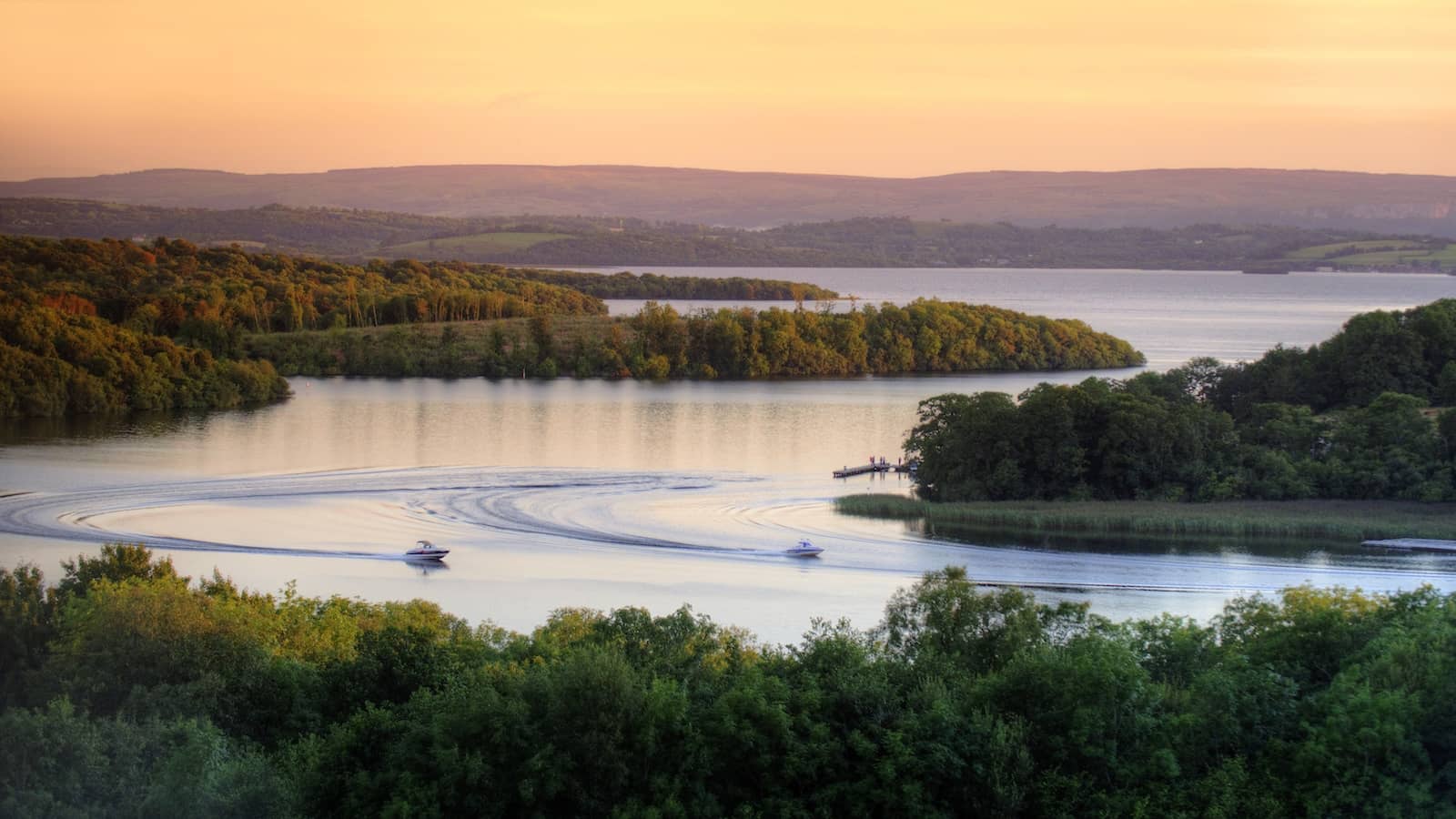 Boats leave gentle wakes across Lower Lough Erne at sunset, surrounded by wooded islands, County Fermanagh.