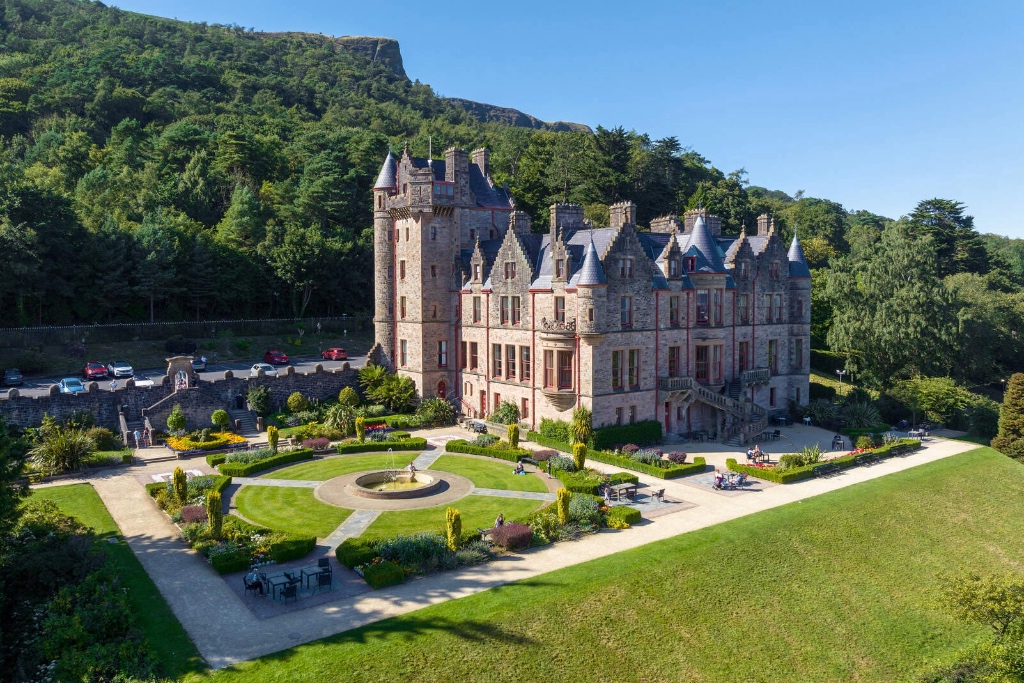 A grand Victorian castle with manicured gardens at the edge of a forested hillside.