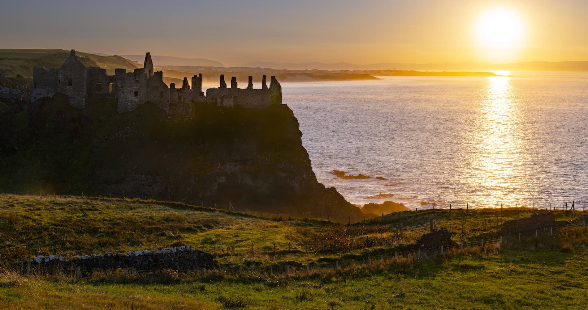 Sunset over Dunluce Castle ruins on the Causeway Coast, with golden light reflecting across the Atlantic Ocean.