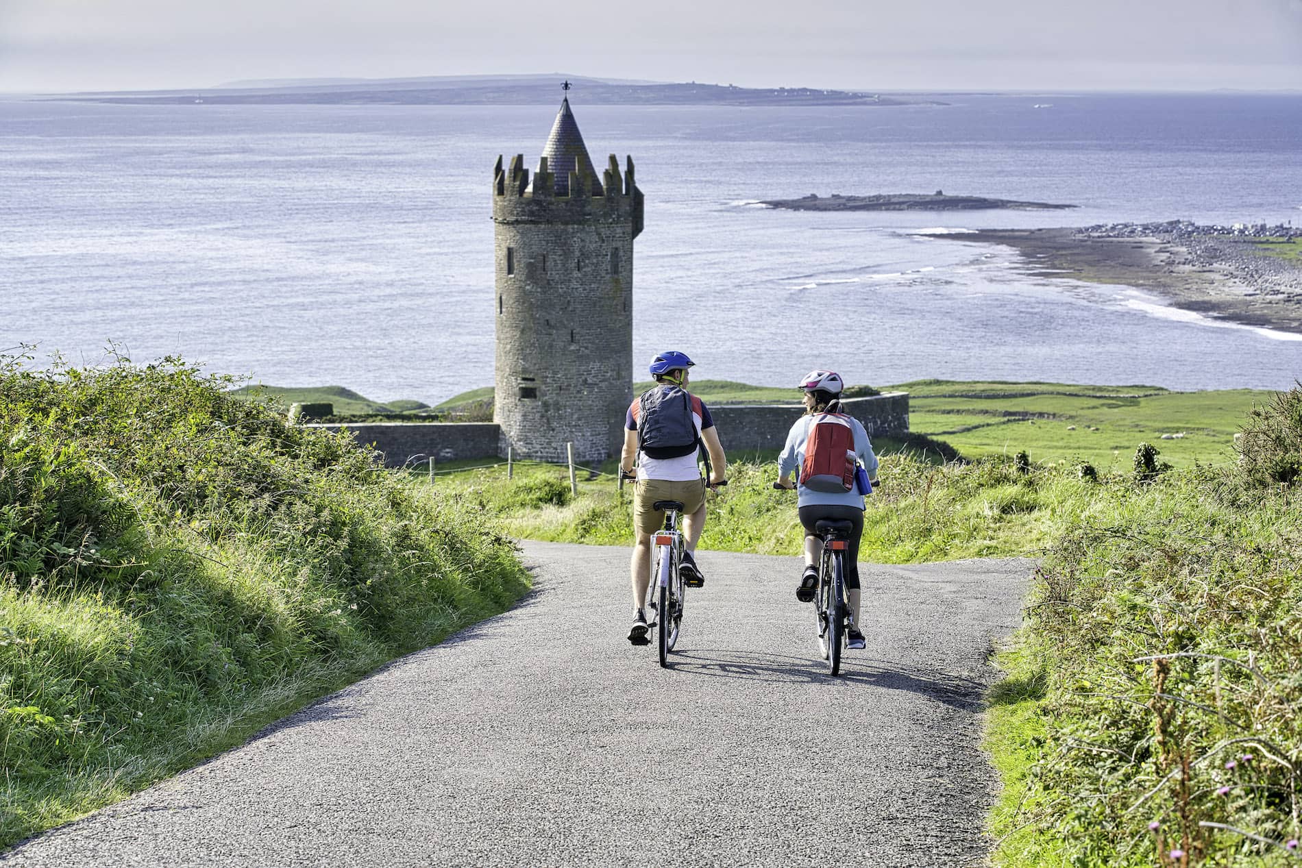 Two cyclists approach Doonagore Castle near Doolin, County Clare, with views of the Atlantic Ocean beyond.