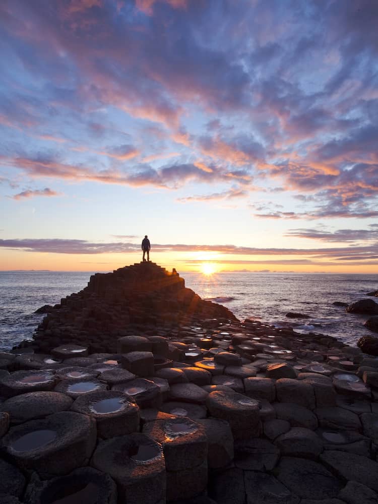 Person standing on Giant’s Causeway basalt columns at sunset, County Antrim.
