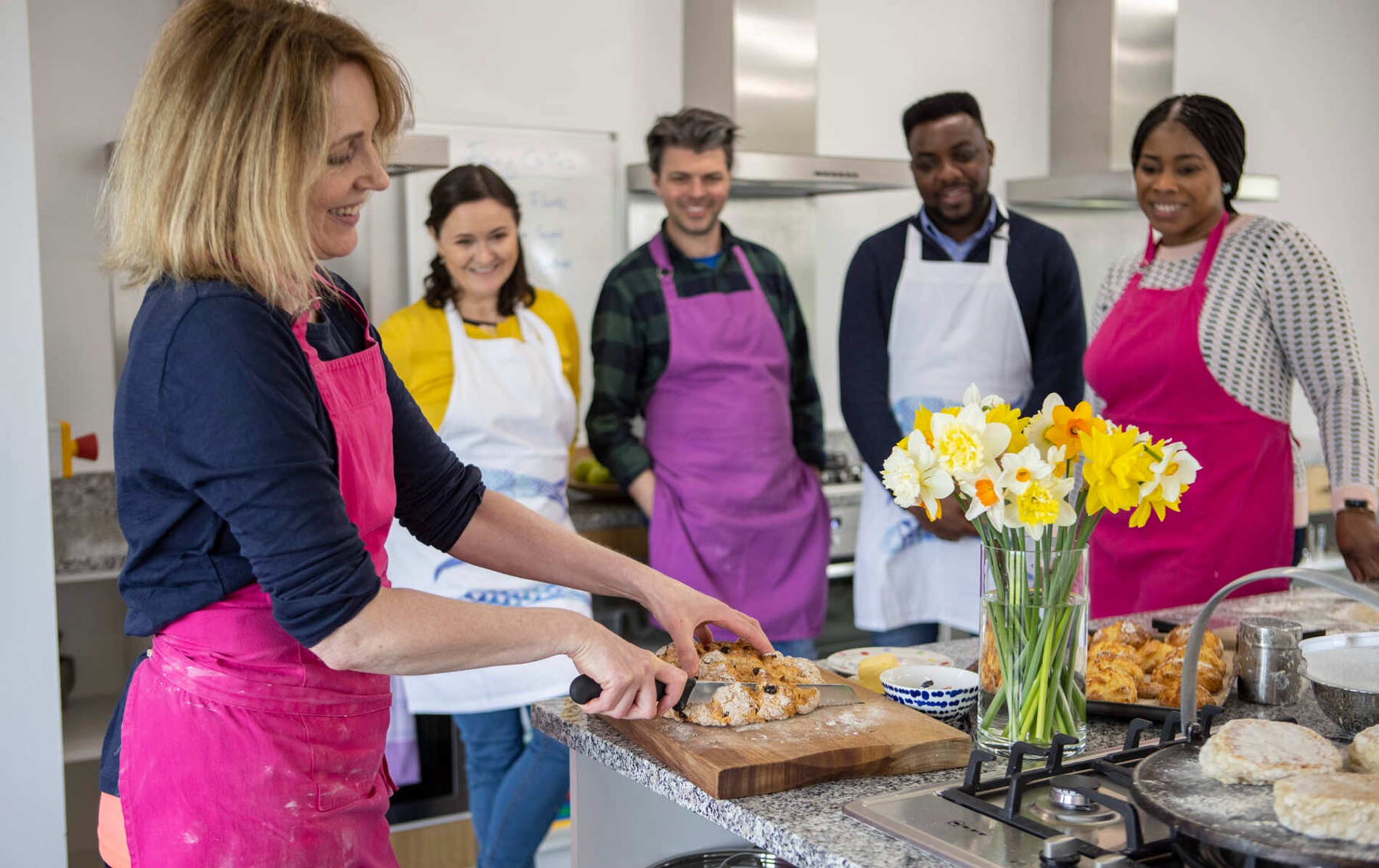 Instructor slices scones as group in aprons smiles at Wee Buns Cookery School in County Tyrone.