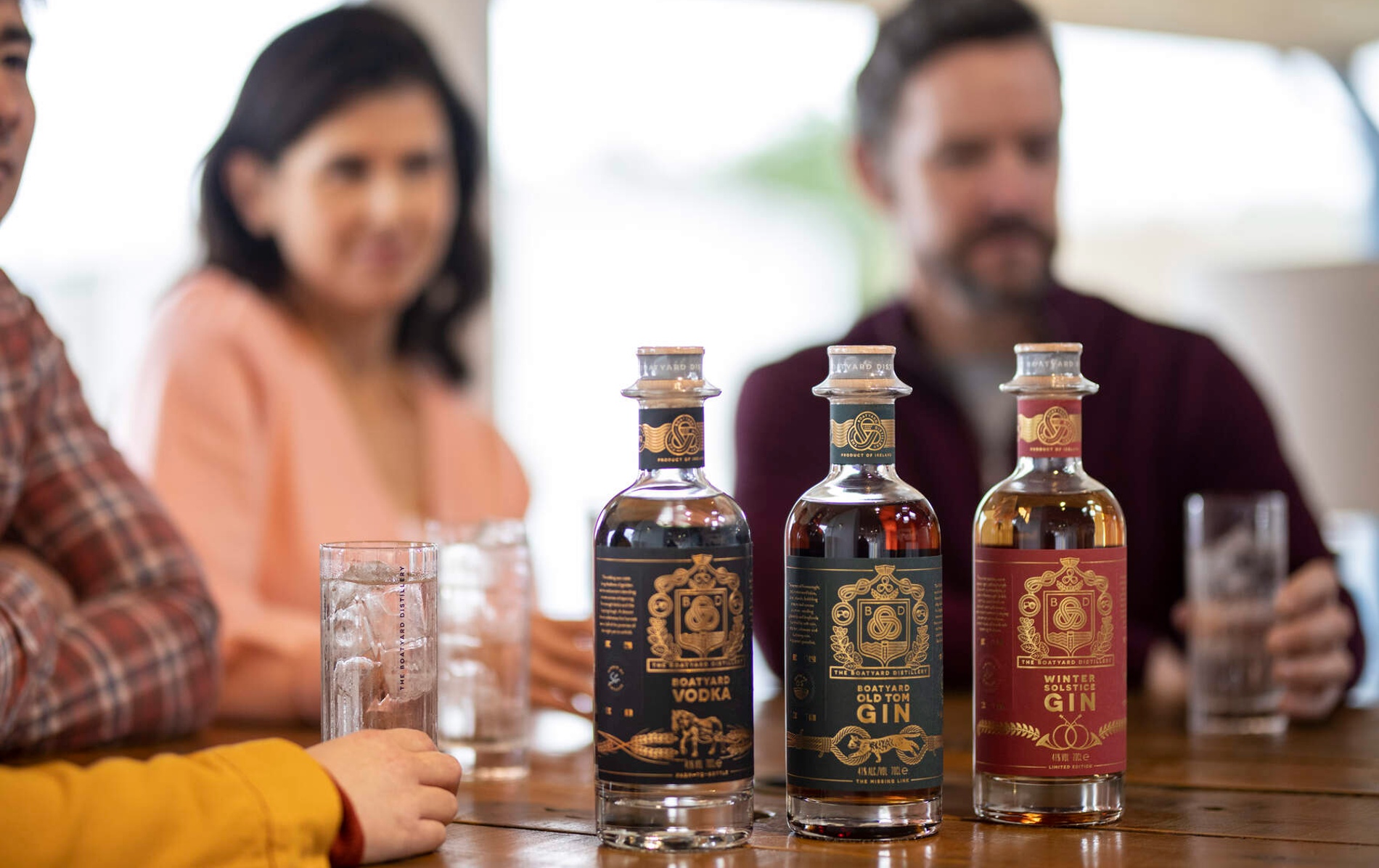 Bottles of Boatyard gin and vodka on a wooden table as visitors sip drinks in a bright tasting room.
